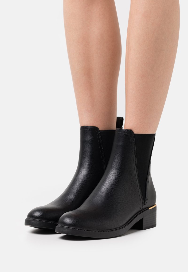 Des bottines noires en cuir présentent une texture lisse, un bout arrondi, des panneaux latéraux élastiques et un petit talon carré avec un accent doré.