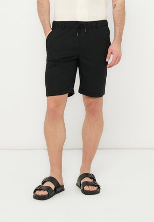Mann iført svarte shorts med snor og svarte sandaler med dobbel stropp, stående mot en ensfarget hvit bakgrunn.
