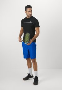 Μαύρο αθλητικό μπλουζάκι με λευκή ρίγα, συνδυασμένο με μπλε σορτς με πλευρικά αξεσουάρ. Μοντέλο που κρατάει μια ρακέτα τένις και φοράει μαύρα παπούτσια.