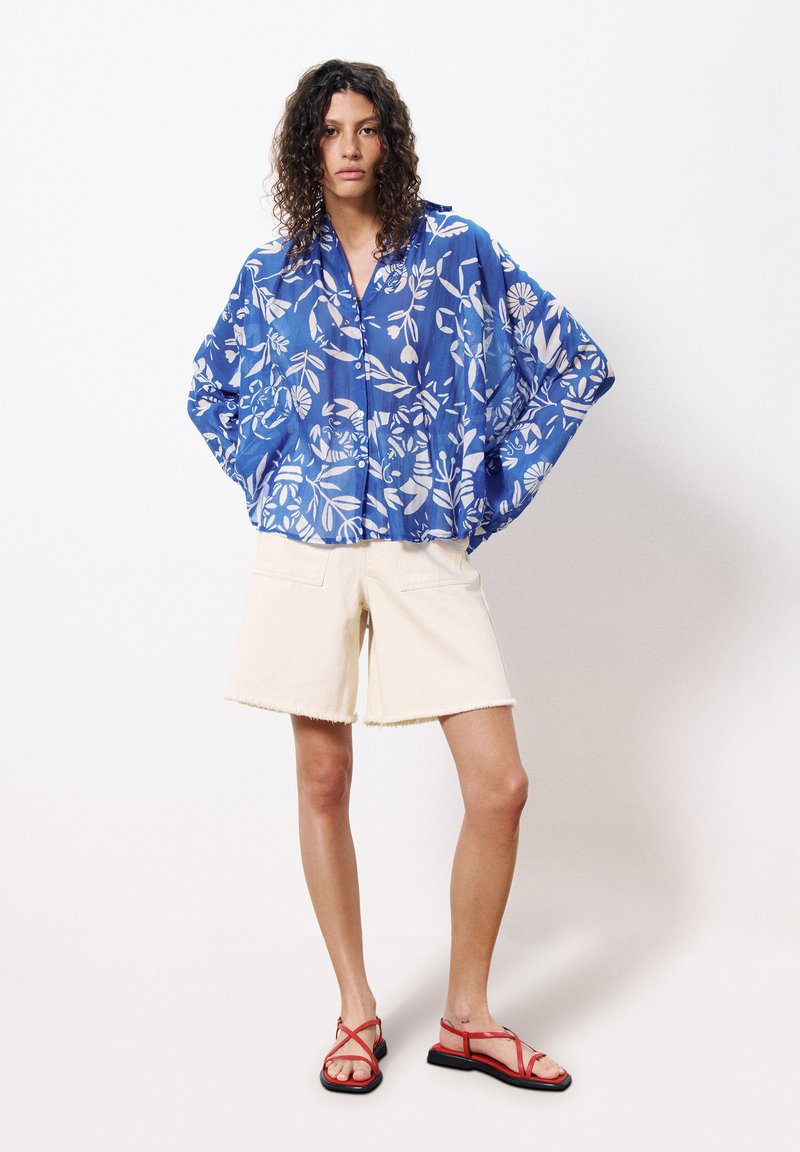 Hellblaues, blumig gemustertes Oversized-Hemd aus leichtem Stoff, kombiniert mit cremefarbenen, verwaschenen Shorts und roten, Riemchensandalen.