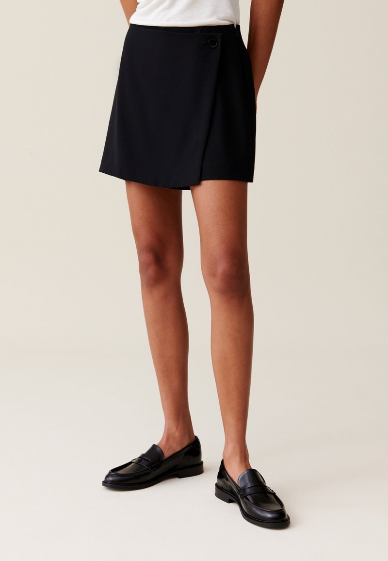 Jupe noire à enfiler avec une fermeture à bouton sur le côté, présentant une texture lisse et une coupe droite, assortie à des mocassins noirs brillants.