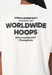 Balta kokvilnas T-kreklu ar uzrakstu "ADIDAS BASKETBALL Izveidots 1949 PASAULS HOOPS Starpkontinentālie čempioni" uz muguras pamanāmā melnā krāsā.