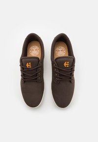Ténis de lona castanhos com atacadores redondos, palmilhas em tom de pele, solas de borracha brancas e um logótipo laranja de lado. Textura suave e design casual.
