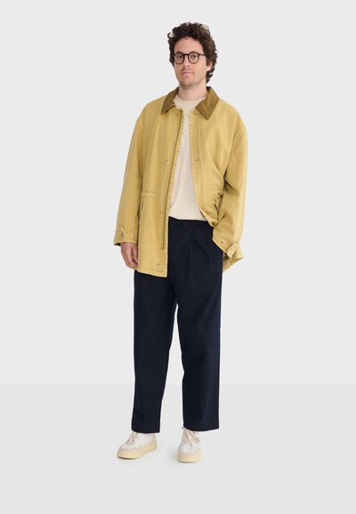 Uomo con capelli ricci e occhiali che indossa una giacca beige, camicia bianca, pantaloni scuri e sneakers bianche, in piedi davanti a uno sfondo neutro.