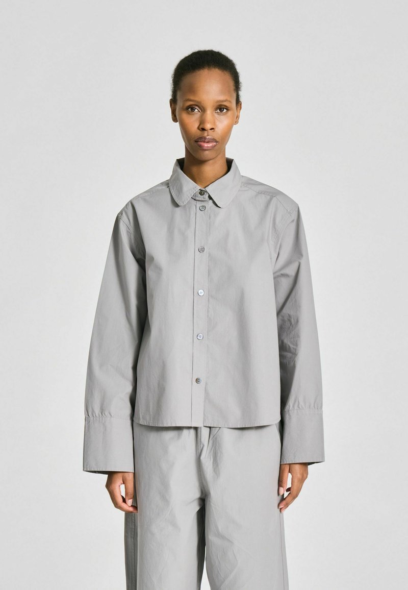 Femme debout devant un fond uni, portant une chemise ample gris clair boutonnée et un pantalon large assorti.