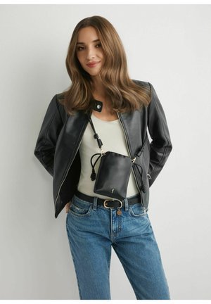 Młoda kobieta w czarnej skórzanej kurtce, białym topie, niebieskich dżinsach i czarnej skórzanej torbie przewieszonej przez ramię, stojąca na tle jednolitego, jasnego tła.