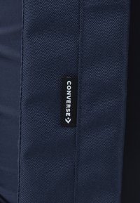 Sac en tissu bleu marine présentant une texture tissée avec une étiquette de logo Converse noire sur le côté, conçu pour la durabilité et le style.