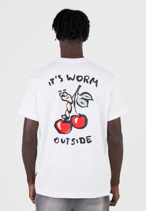 Personne portant un T-shirt blanc avec un ver de dessin animé sur des cerises et le texte « IT'S WORM OUTSIDE » au dos, debout devant un fond uni.