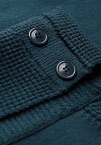 Stickad tealtyg med en strukturerad mönster, med två svarta knappar fästa vid kanten. Mjuk och varm känsla.