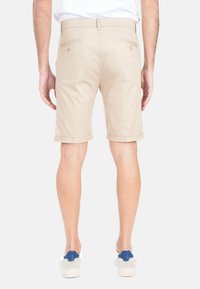 Pantaloni corti beige in cotone con risvolti, dotati di tasche posteriori con bottone e una trama liscia. Indossati con sneaker bianche e blu.