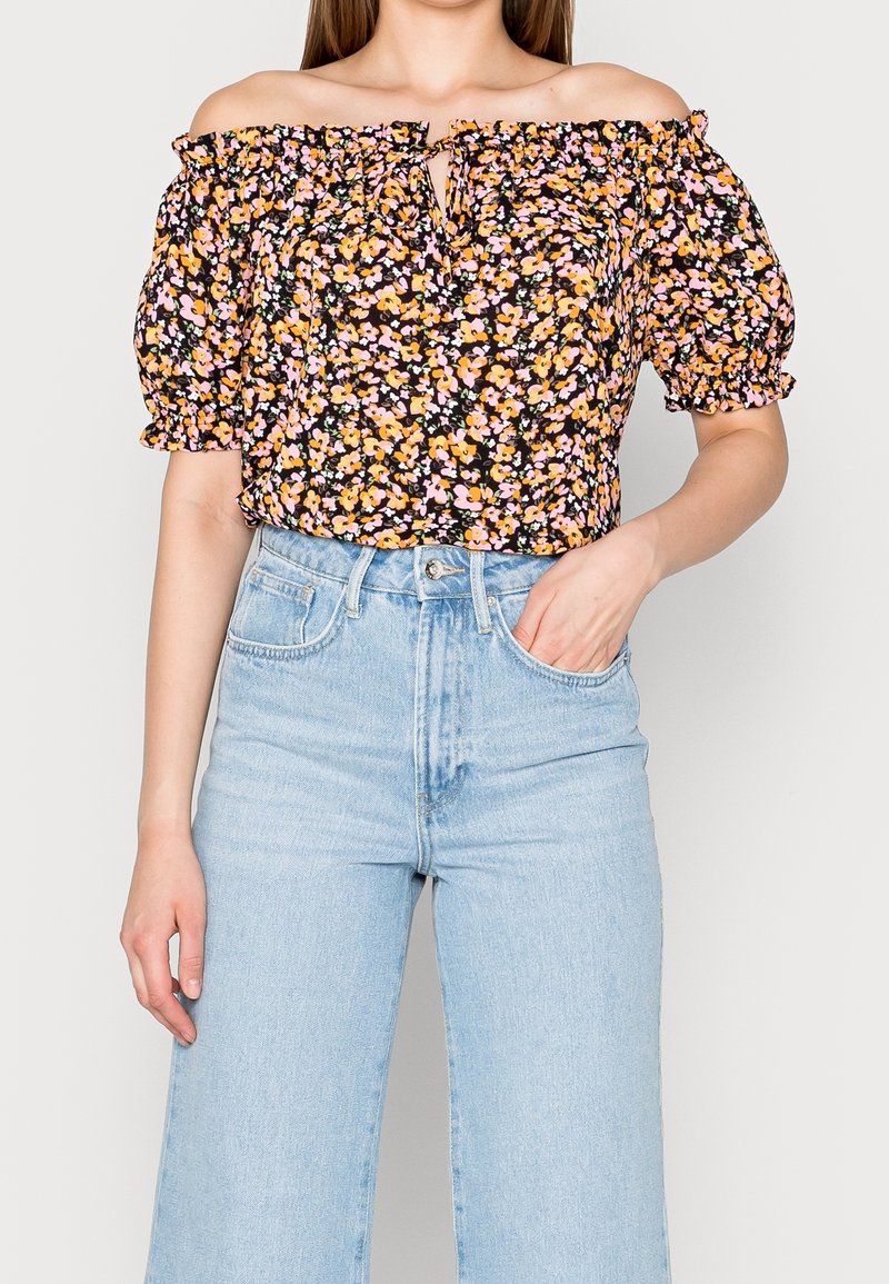 Blouse fleurie à épaules dénudées avec un col élastique, des manches courtes bouffantes, base noire, et fleurs multicolores roses et orange. Associée à un jean taille haute bleu clair.