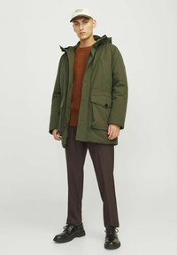 Chaqueta acolchada verde oliva con capucha, cierre de cremallera y bolsillos delanteros, combinada con un suéter texturizado color óxido y pantalones marrones, rematada con una gorra beige.