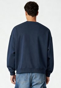 Homme portant un sweat-shirt à col rond bleu marine et un short cargo en denim bleu clair, debout de dos sur un fond uni.