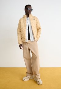Quiltad beige jacka med ståkrage, kombinerad med en randig skjorta med knappar och ljusbeiga vida byxor. Skorna är klumpiga krämfärgade träskor.