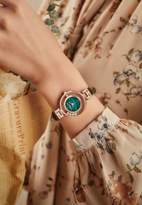 Reloj de pulsera en oro rosa con una esfera circular, dial de malaquita verde, tres acentos de diamante y una correa negra. Rodeado de tela floral.