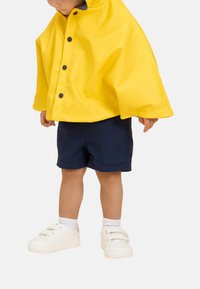 Gelber Regenmantel mit schwarzen Knöpfen, marineblaue Shorts und weiße Sneakers. Glatte, wasserdichte Textur mit hohem Kragen und kurzen Ärmeln.