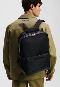 Mochila negra con tela lisa, cierre con cremallera y acentos texturizados. Presenta un diseño moderno y correas de hombro ajustables.