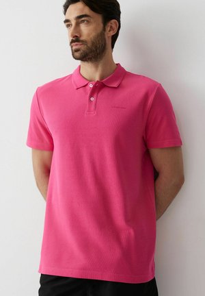 Mężczyzna z brodą ubrany w jaskraworóżową krótką koszulkę polo stoi przy białej ścianie i patrzy w lewo.