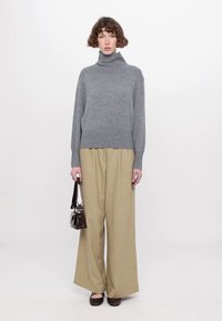 Grå turtleneck genser, avslappet passform, parret med vide beige bukser. Modellen holder en liten mørk veske, står foran en lys bakgrunn.