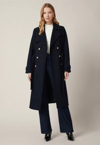 Granatowy płaszcz dwurzędowy z złotymi guzikami, szerokimi klapami i mankietami z paskiem, noszony na białej bluzce i ciemnych dżinsach. Gładka tekstura materiału.