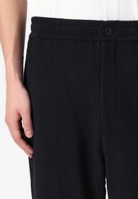 Svarta ribbade byxor med en texturerad yta, utrustade med ett brett elastiskt midjeband och en enda knappstängning. Handen placerad vid sidan.