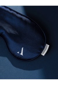 Granatowa satynowa maska na oczy o gładkiej fakturze, z białym logo i przymocowaną etykietą z jednej strony, spoczywająca na ciemniejszym niebieskim tle.