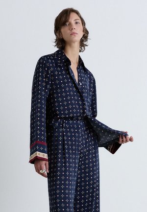 Jeune personne portant une chemise à manches longues bleu marine à motifs et un pantalon assorti, tenant l’ourlet de la chemise devant un fond clair uni.