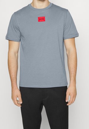 Graues Baumwoll-T-Shirt mit kurzen Ärmeln, mit einem roten rechteckigen Patch mit dem "HUGO"-Logo auf der Brust. Glatte Textur, klassische Passform.