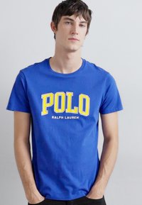 Camiseta de algodón azul con un gran logo amarillo "POLO", mangas cortas, cuello redondo y una textura suave. Diseñada por Ralph Lauren.