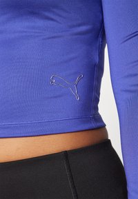 Long-sleeve purple athletic shirt made of smooth fabric with a fitted design. Features a silver Puma logo at the waist. Paired with black leggings.