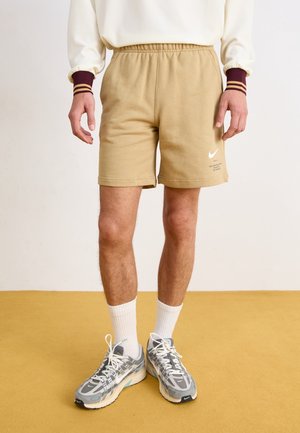 Homme portant un short beige Nike, une chemise blanche à manches longues avec des poignets bordeaux, des chaussettes blanches et des baskets grises, debout sur un sol jaune.
