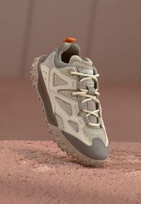 Hellgrauer Sportschuh mit Mesh-Obermaterial, Gummielementen und strukturiertem Profil. Hervorstechendes Detail ist das farbenfrohe orangefarbene Fersentäfelchen und die neutralen Schnürsenkel.