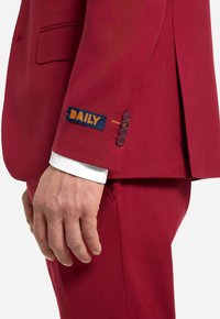 Giacca rossa con etichetta blu con la scritta "DAILY," cinque bottoni e un polsino di camicia bianca visibile sotto. Tessuto dalla texture liscia.