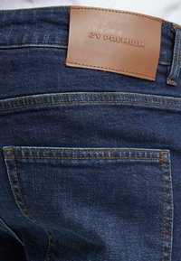 Jean de mezclilla azul oscuro con una etiqueta de cuero que dice "2Y PREMIUM" en la parte posterior. Bolsillos cosidos y detalle de hilo naranja en contraste.