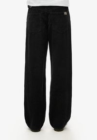 Zwarte corduroy broek met een wijde pijpdesign, voorzien van twee achterzakken en een klein merklabel aan de rechterkant boven de taille.