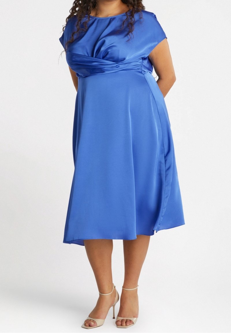 Robe en satin bleue à manches courtes, avec un détail torsadé au niveau du corsage et une jupe évasée en ligne A, accompagnée de talons à bride cheville.
