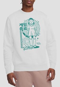 White sweatshirt featuring a green graphic of a girl in a dress with the text "Red Light Green Light" and elements related to a game theme.