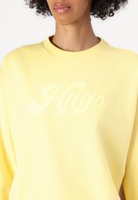 Felpa gialla realizzata in un morbido tessuto con collo rotondo e vestibilità oversize, caratterizzata da un logo "Hugo" impresso sul davanti.