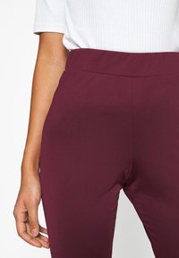 Burgundy stretchy leggings with a smooth texture; high waistband and fitted design, partially shown with a white ribbed shirt.