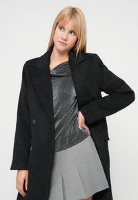 Palton negru din lână cu reveruri largi și nasturi dubli, purtat peste un top argintiu metalic și o fustă plisată gri. Textură netedă, croială cambrată.