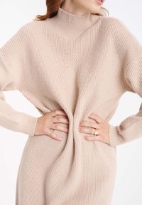 Pull en tricot côtelé beige avec un col montant et une coupe ample. Présente un design torsadé à la taille et des manches longues. Texture douce.