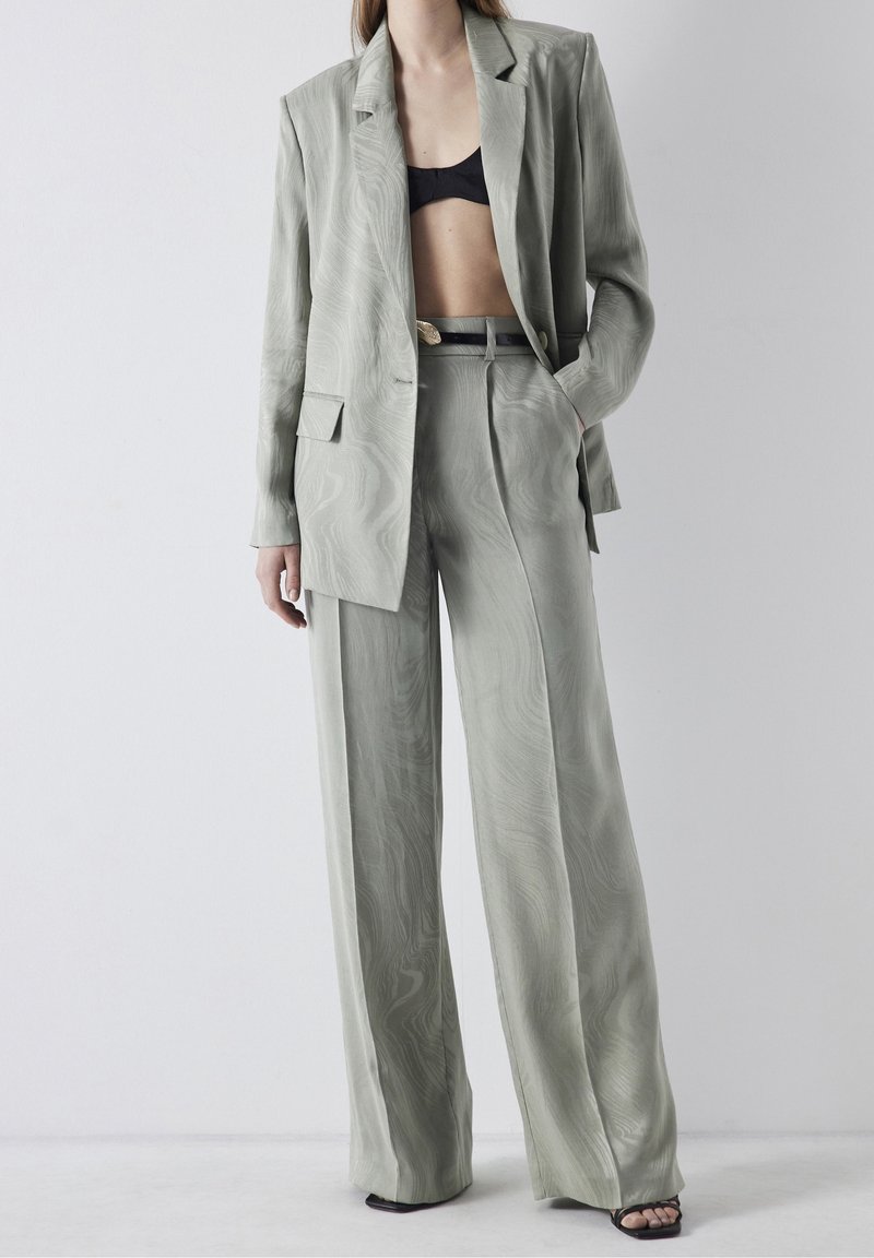 Femme portant un pantalon large texturé gris clair et un blazer oversize assorti avec un soutien-gorge noir et des talons ouverts, debout devant un fond blanc uni.