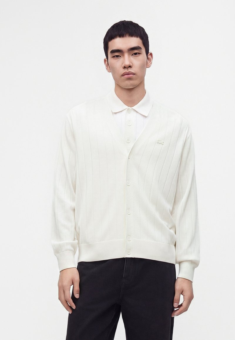 Jeune homme portant un cardigan blanc boutonné sur une chemise blanche à col et un pantalon noir sur un fond blanc uni.