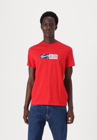Rød bomulds T-shirt med korte ærmer, udstyret med et logo grafik i blå og hvid på fronten. Afslappet pasform med rund halsudskæring.