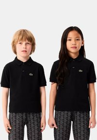 Czarne koszulki polo z krótkim rękawem, z zielonym haftowanym logo, zestawione z wzorzystymi czarnymi spodniami. Materiał wydaje się gładki i jednolity.