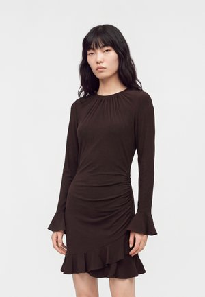 Jeune femme aux longs cheveux noirs portant une robe marron foncé cintrée avec manches longues, poignets volantés et un ourlet volanté sur un fond uni.