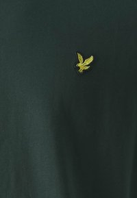 T-shirt verde scuro con un design aderente, caratterizzato da un piccolo logo di un'aquila gialla sul lato sinistro del petto, realizzato in morbido cotone.