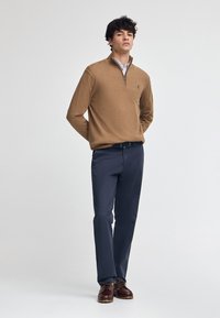 Brauner Reißverschluss-Pullover über einem Hemd mit Kragen, kombiniert mit blauen Hosen und braunen Schuhen, mit einem lockeren Sitz und glatter Textur.