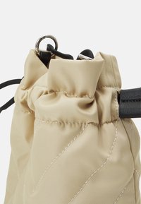 Borsa beige trapuntata con parte superiore arricciata, spallacci neri regolabili e dettagli in metallo argentato. Texture liscia con motivi di cucitura sottili.