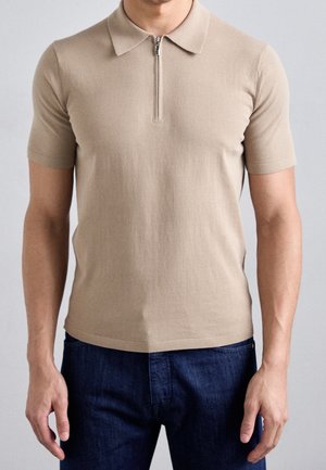 Homme portant un polo beige à manches courtes avec fermeture éclair sur le devant et un jean bleu foncé sur un fond gris uni.